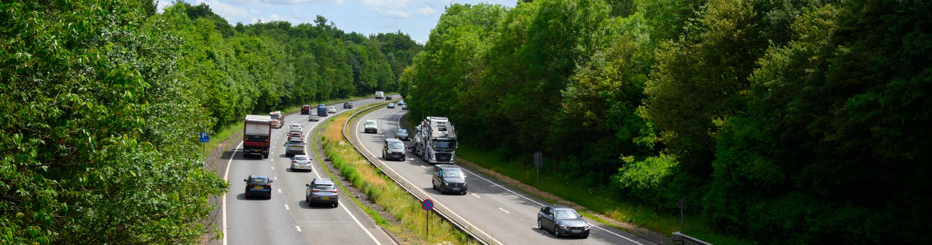 A dual carriageway in England with vehicles driving on it. Tall green trees line the side of the dual carriageway.
