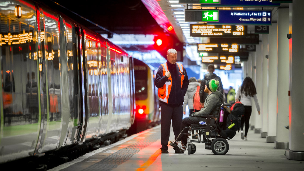 railway passenger using a wheelchair speaks to rail member of staff on a station platform with a train on the left of the image