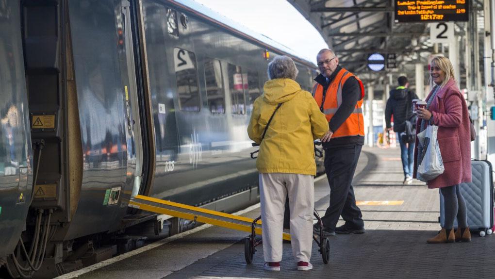 Staff member assisting passenger with mobility aid onto train with ramp deployed at a train station