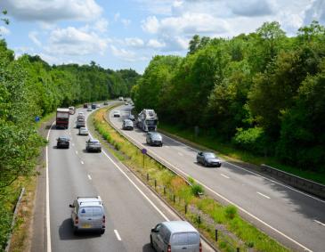 A dual carriageway in England with vehicles driving on it. Tall green trees line the side of the dual carriageway.